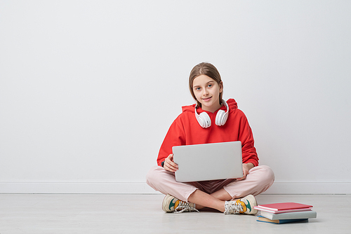 Portrait of content modern high school girl in red hoodie sitting with crossed legs on floor and using portable computer