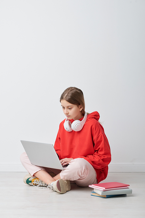 Curious schoolgirl with wireless headphones around neck performing online task on laptop