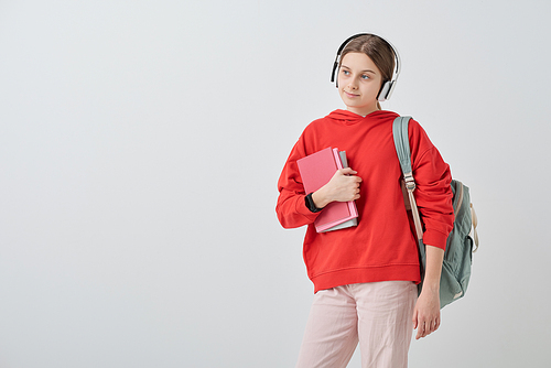 Teenage girl in red hoodie and white jeans holding books by chest and listening to her favorite music in headphones in isolation