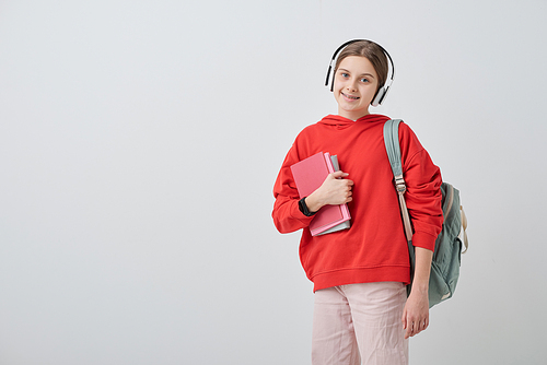 Portrait of smiling pretty teenager in headphones wearing satchel and holding school books