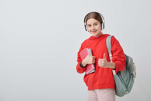 Pretty cheerful girl with rucksack on back and books in hand showing thumb up while listening to music in headphones in isolation