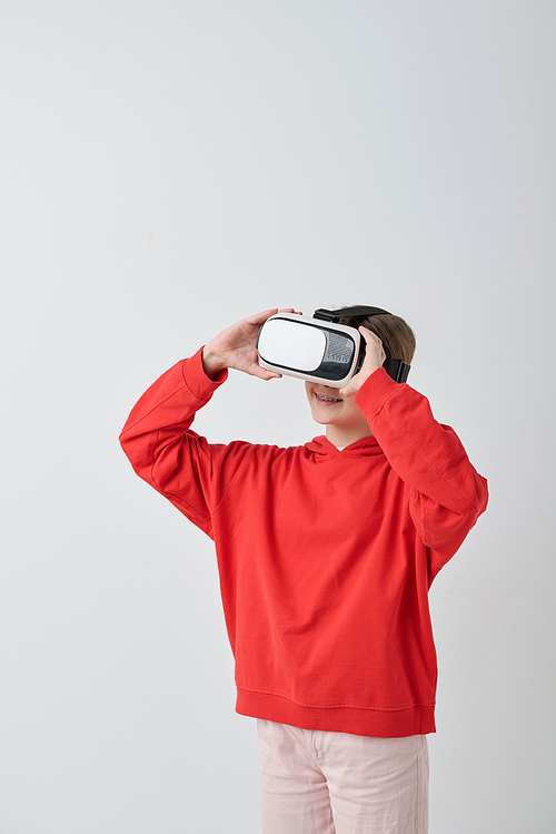 Cheerful modern teenager in casualwear watching video in vr headset while standing against white background in isolation