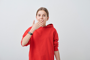 Amazed teenage girl in red hoodie and smartwatch keeping one hand by open mouth while standing in front of camera in isolation
