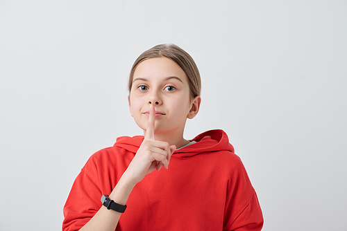 Portrait of teenage girl in red hoodie holding finger near lips while making silence sign against white background