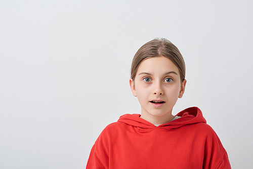 Scared or surprised teenage girl in red hoodie standing in front of camera against white background and looking at you