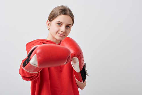 Confident and strong teenage girl in red activewear and boxing gloves doing kick while exercising in front of camera in isolation