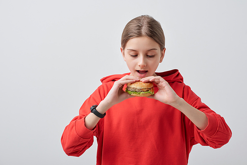 Youthful girl in red hoodie holding appetizing hamburger by her open mouth while going to eat it against white background in isolation