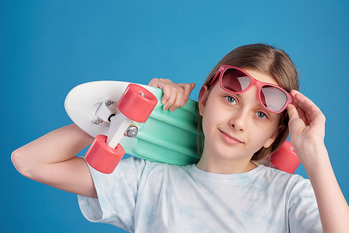 Portrait of content teenage skateboarder holding skateboard on shoulder and adjusting sunglasses