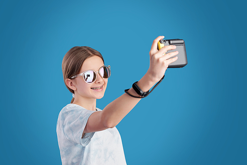 Smiling teenage girl in sunglasses and t-shirt holding photocamera in front of herself while making selfie against blue background