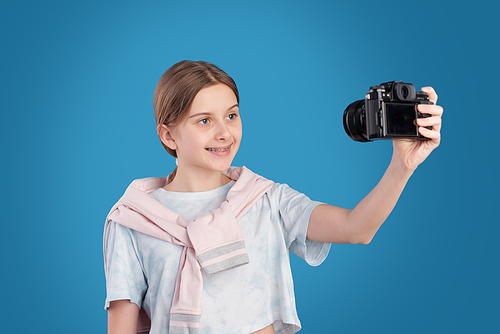 Happy teenage girl in white t-shirt standing against blue background and photographing herself on photocamera in isolation