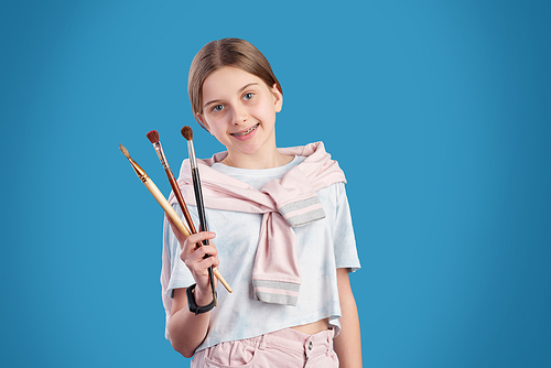 Happy youthful girl in casualwear showing you collection of paintbrushes for professional painting while standing over blue background
