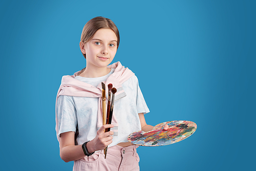 Portrait of art school student posing with paintbrushes and palette against blue background