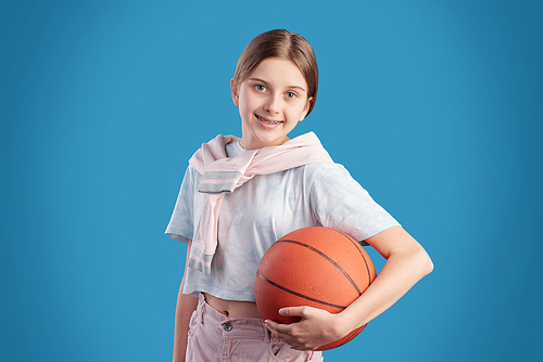 Cheerful active teenage girl in t-shirt holding ball for playing basketball while standing in front of camera against blue background