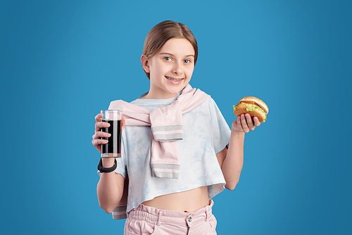 Portrait of positive teenage girl eating unhealthy hamburger and drinking cola against blue background