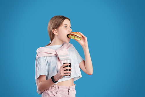 Teenager with sweater wrapped around shoulders eating burger and drinking cola against blue background