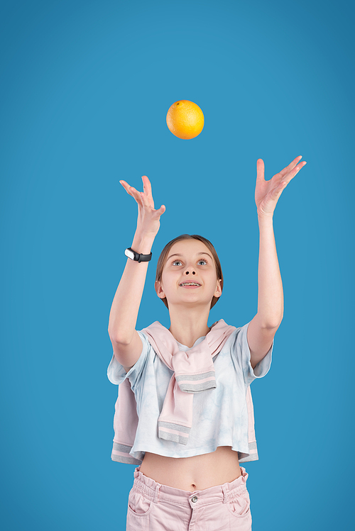Energetic teenage girl throwing fresh orange while playing with it in front of camera against blue background in isolation