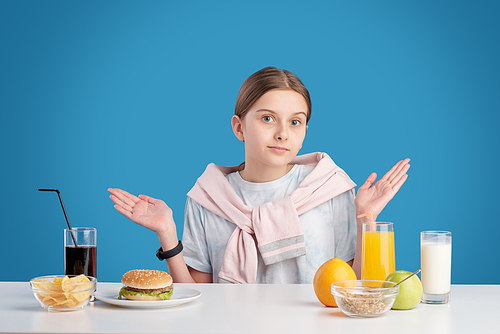 Portrait of confused teenage girl shrugging shoulders while choosing between healthy and unhealthy food