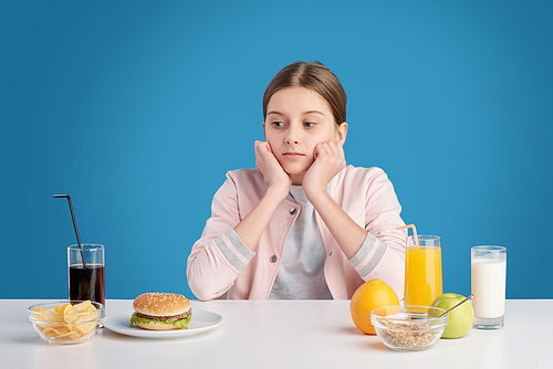 Teenage girl choosing between fresh organic healthy eating and junk food while sitting by table in front of camera in isolation