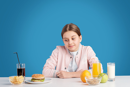 Teenage girl sitting at table and looking at unhealthy burger and cola while facing with difficult choice of nutrition