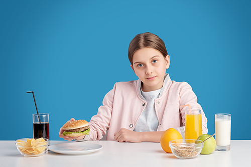 Pretty teenage girl looking at you while sitting by table and going to eat hamburger, potato chips and drink cola for lunch