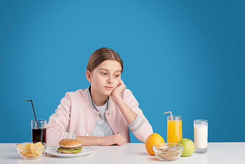 Thoughtful female teenager sitting at table with healthy and unhealthy food and choosing nutrition