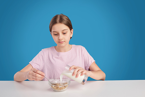 Teenage girl sitting at table and adding milk into cereal while having breakfast, blue background