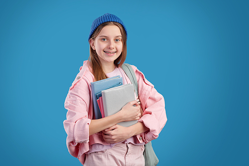 Portrait of smiling attractive girl with braces standing against blue background and holding heap of textbooks