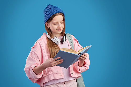 Content schoolgirl with headphones around neck standing against blue background and reading textbook