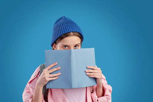 Shy teenage girl in blue beanie and pink denim jacket peeking out of open book by her face while standing in front of camera