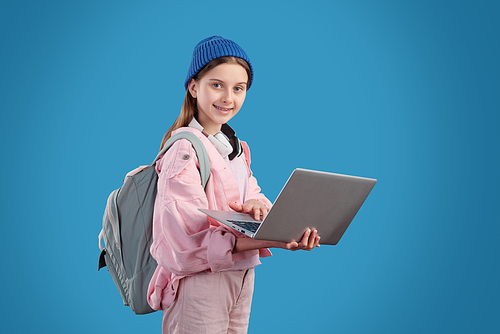 Happy teenage girl in casualwear holding laptop and surfing in the net while standing in front of camera over blue background