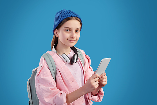 Portrait of content teenage girl in beanie hat wearing satchel using mobile app against blue background