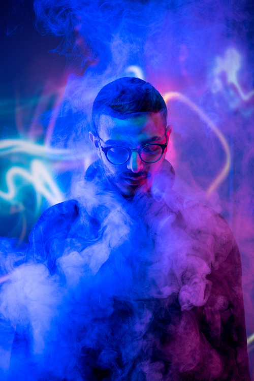 Head of young man in eyeglasses standing in isolation in front of camera among smoke and blue neon light