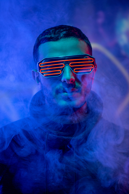 Portrait of serious young mixed race man in neon geometric goggles standing in smoked dark blue room