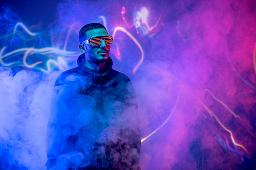 Serious cool guy in neon goggles and sweatshirt standing in blue and pink smoked room