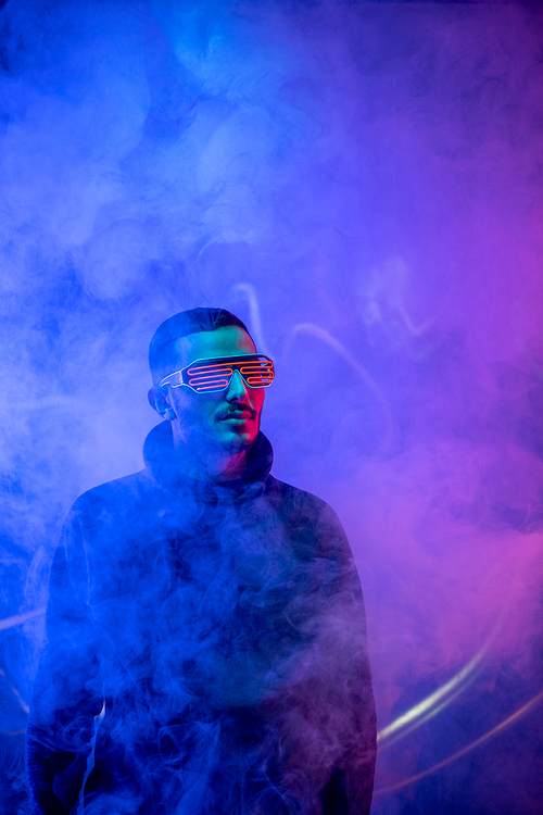Studio shot of bearded young man in red spiral eyeglasses standing in the smoke among blue neon light