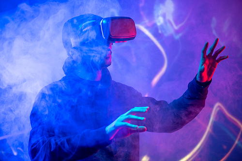 Young Arabian man using virtual reality device gesturing hands in smoked VR room