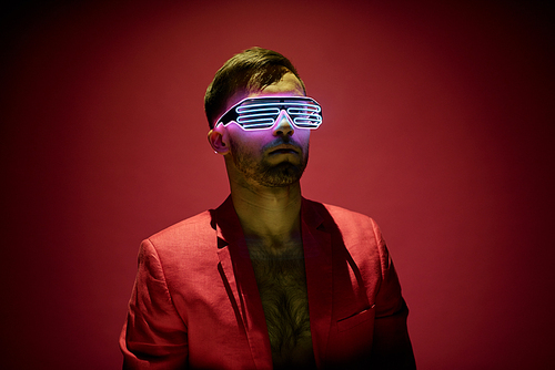 Contemporary young man in red jacket and high tech eyewear standing in darkness against maroon background in isolation