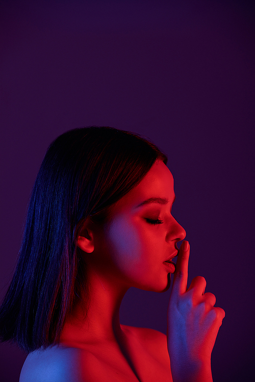 Pretty young brunette woman holding forefinger by her mouth expressing quietness against dark background in isolation