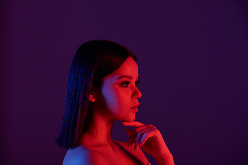 Side view of pensive naked young woman with straight hair touching chin in neon lights