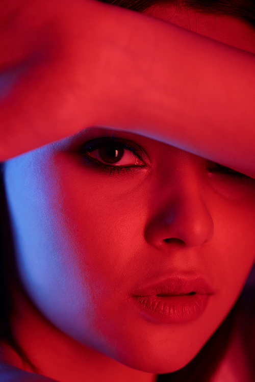 Portrait of serious passionate young woman with smokey eyes covering forehead with arm in neon light