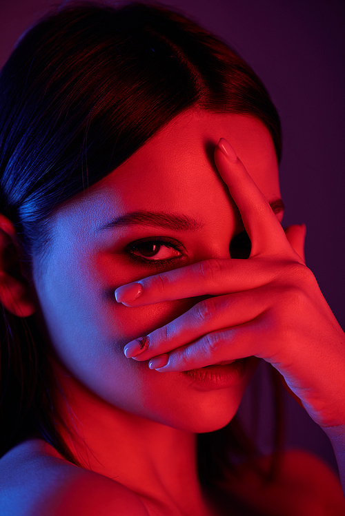 Face of pretty young female with hand by her face peeking through fingers while looking at you over black background