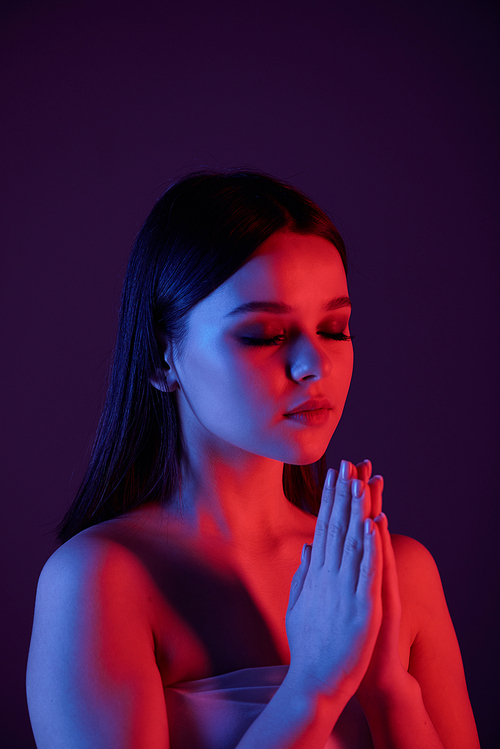 Serene calm young woman with naked shoulders meditating with closed eyes against dark blue background, neon light