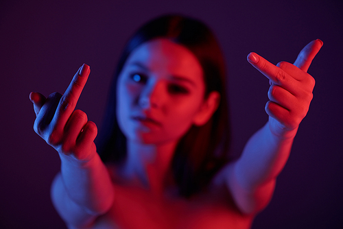 Young rude female showing indicent gesture while standing in darkness and keeping her hands in front of camera