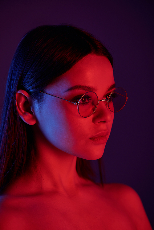 Side view of serious naked woman in round glasses under neon light in dark room