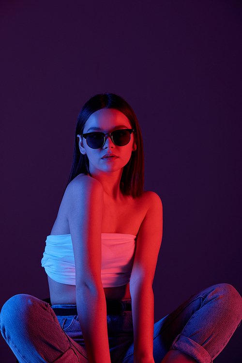 Young stylish woman in sunglasses, white tanktop and blue jeans sitting on the floor of studio over dark background