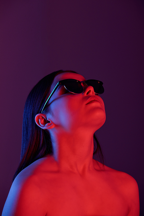 Serious naked young woman in sunglasses looking up under dark neon light