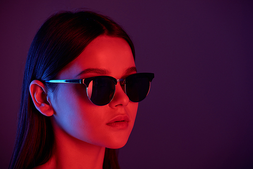 Head of young stylish female with perfect make-up wearing sunglasses in front of camera against dark neon background
