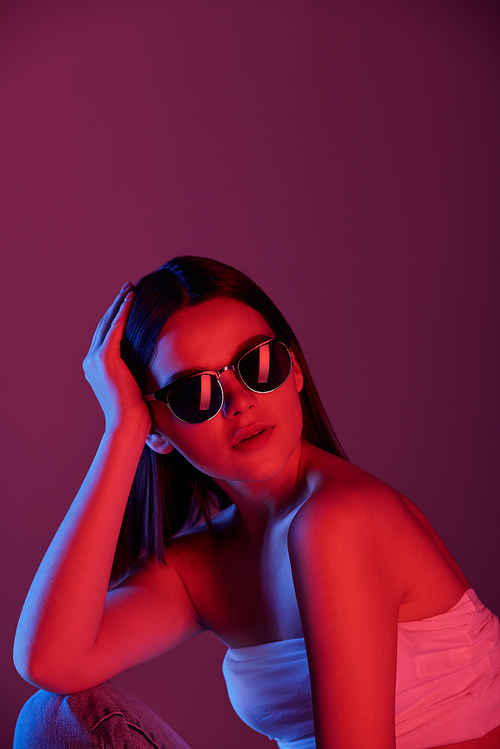 Pretty glamorous girl in sunglasses and white tanktop posing in front of camera against black background in isolation