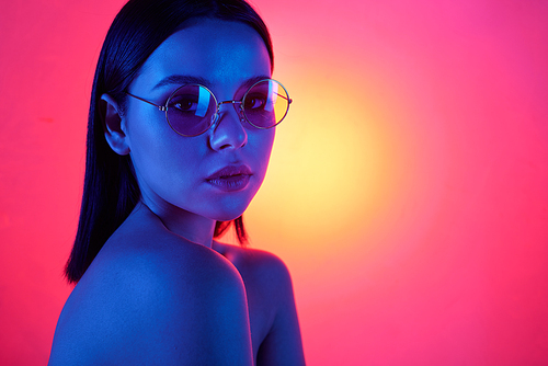 Young brunette female in stylish eyeglasses with perfect smooth hair and natural makeup looking at you on neon pink background