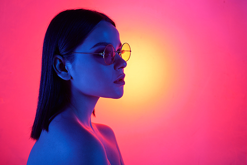 Blue light falling on serious naked girl in stylish round glasses against bright pink background